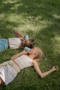 Two girls lie in the grass holding cups to their ears and mouth.