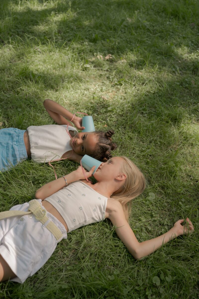 Two girls lie in the grass holding cups to their ears and mouth.