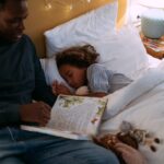 Child sleeps while parent sits aside them with a book in their lap.